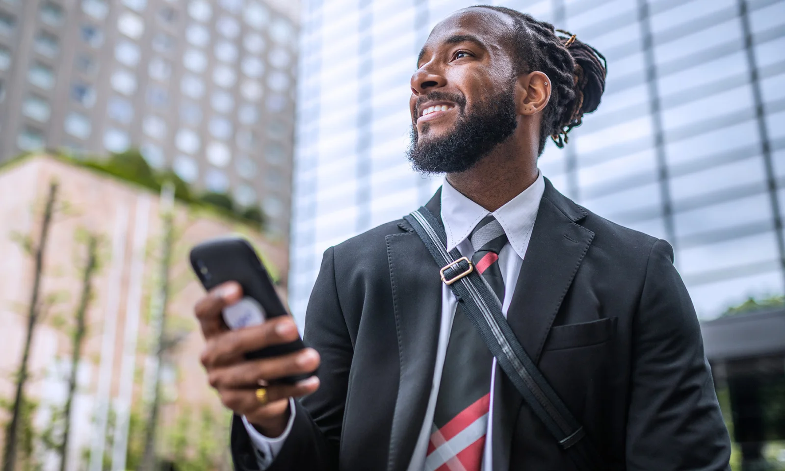 Smiling businessman in a suit holding a smartphone in a city environment with modern buildings in the background.