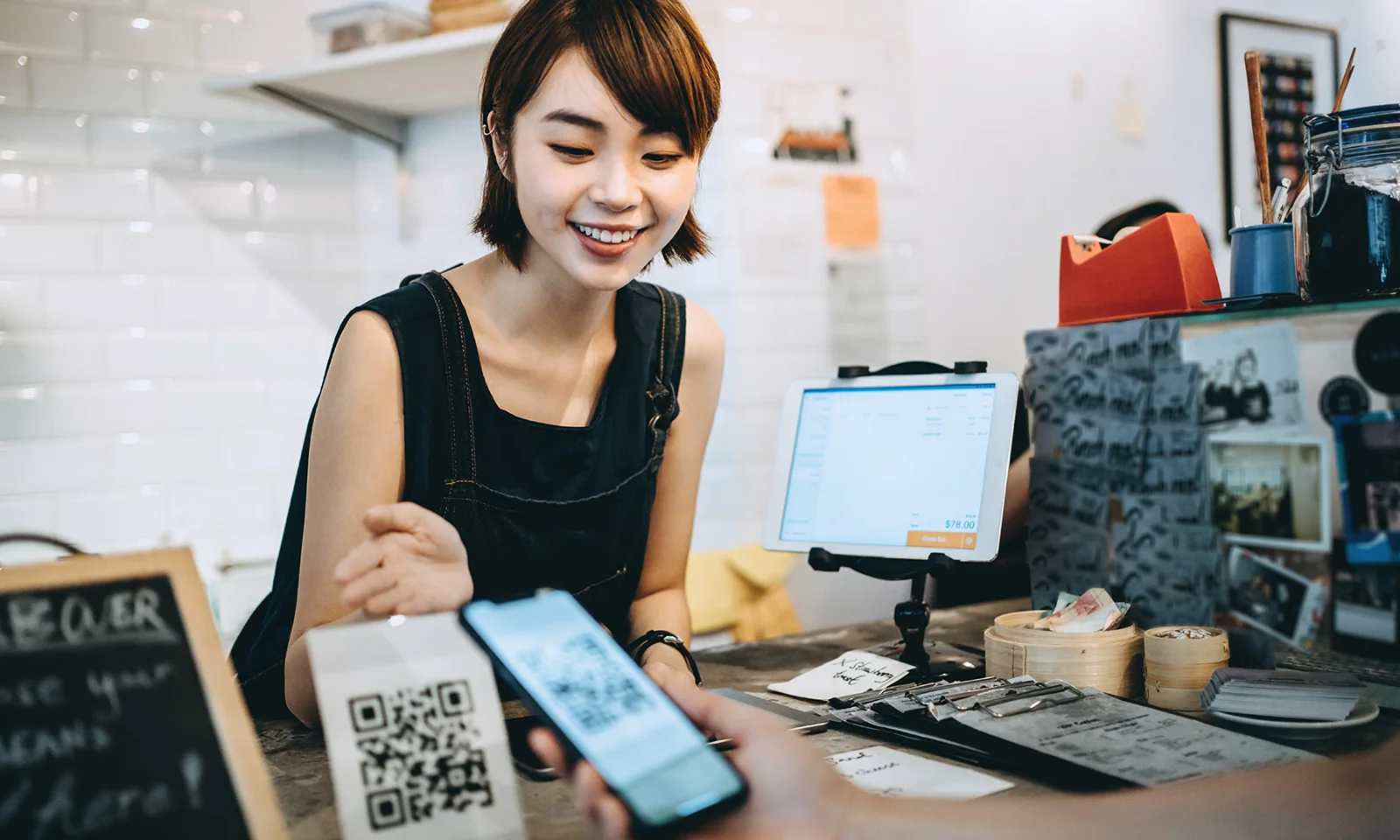 Smiling barista at a café counter accepts a QR code payment from a customer using a smartphone, with a digital register visible in the background.