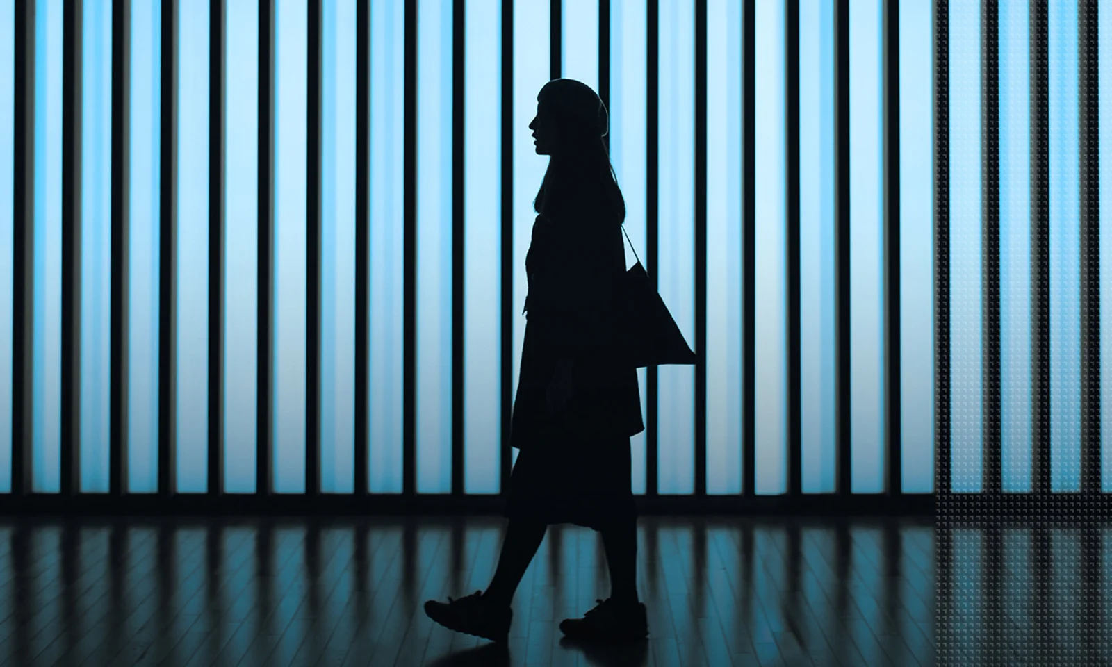 Silhouette of a woman with a bag walking in front of a backlit, striped wall in a modern corridor.