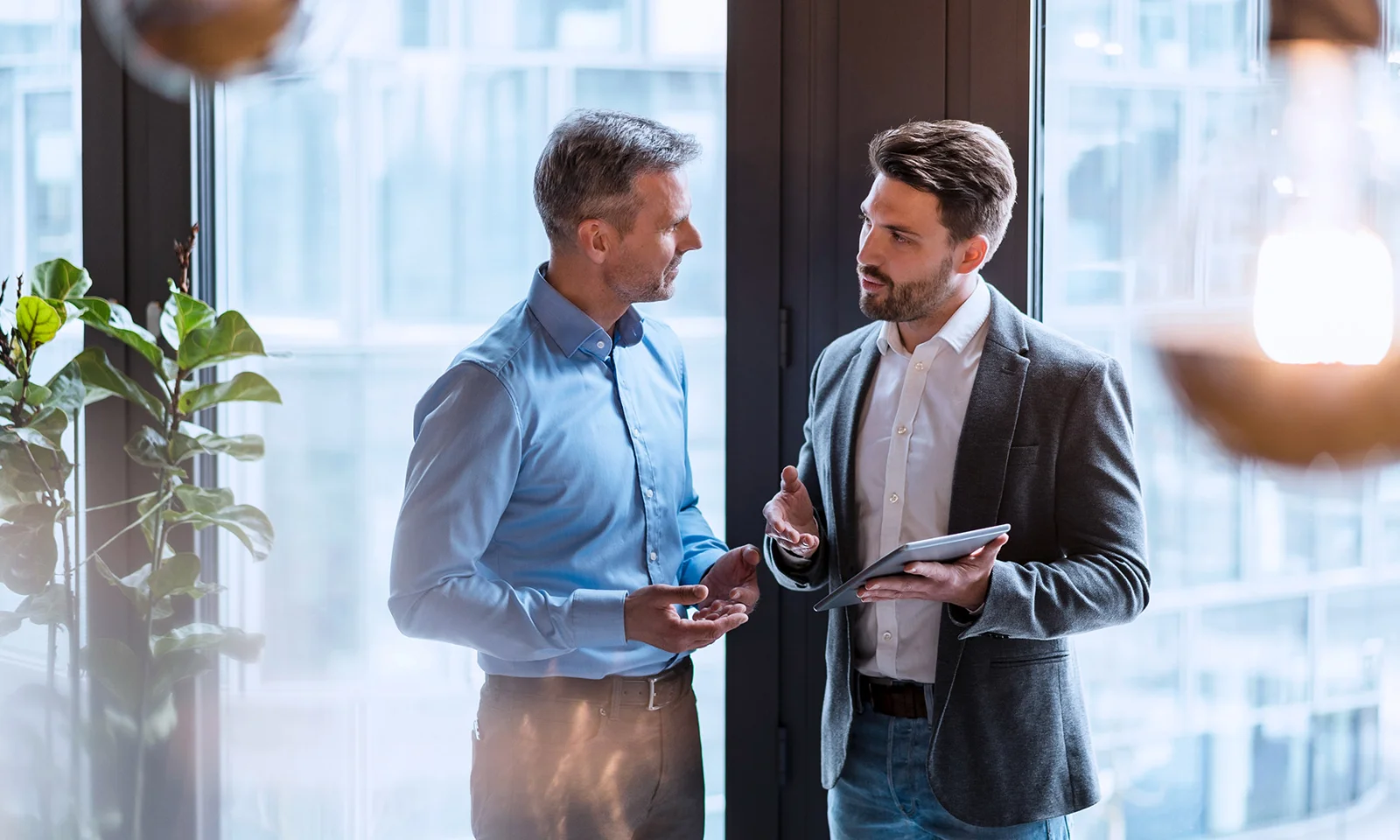 Two businessmen engaged in a discussion, one holding a tablet, in a modern office with large windows and greenery.