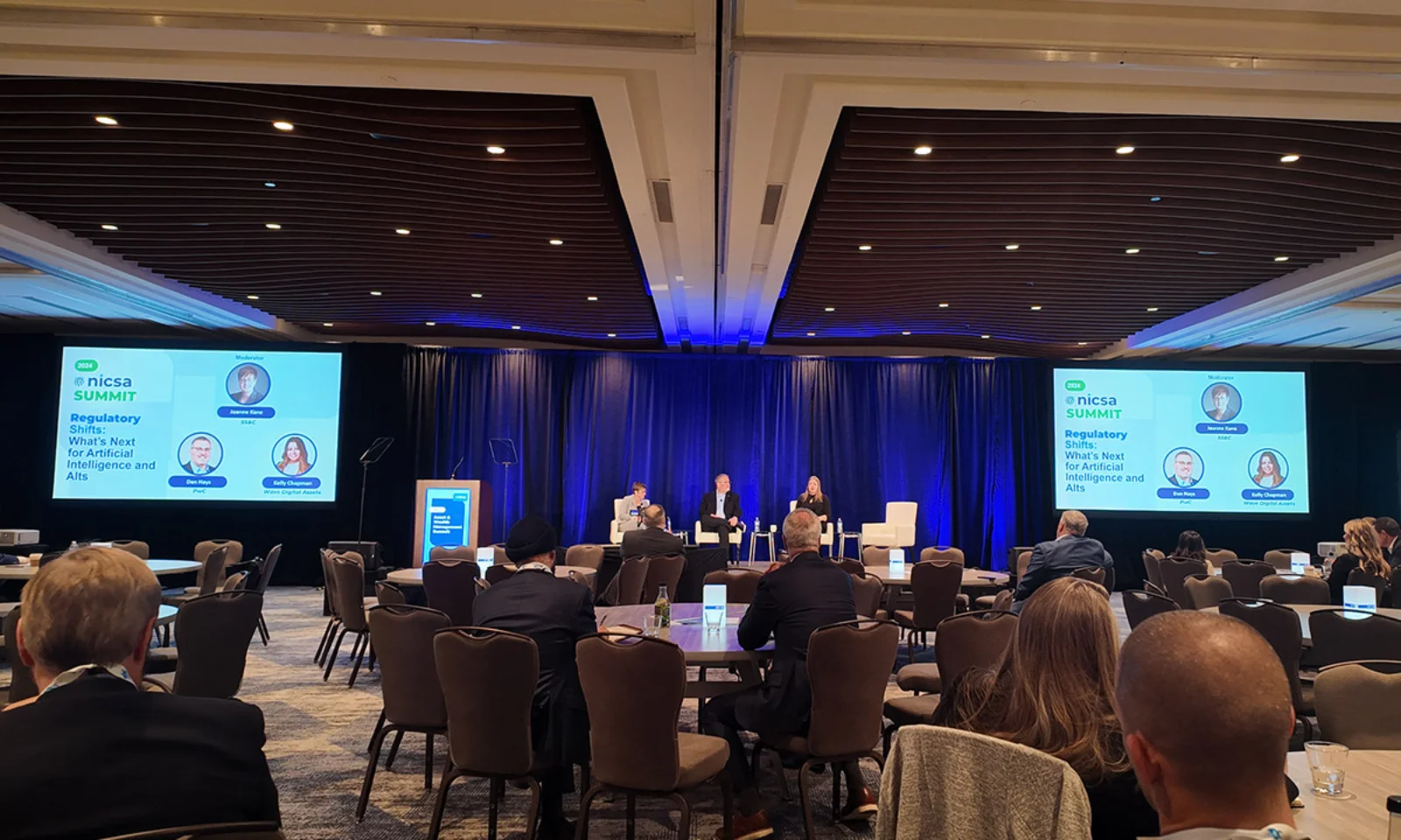 A wide-angle view of the panel discussion at the NICSA Summit 2024, focusing on &quot;Regulatory Shifts: What&#039;s Next for Artificial Intelligence and Alts.&quot; The stage features three panelists and a moderator, with a blue-lit backdrop and an audience in the foreground.