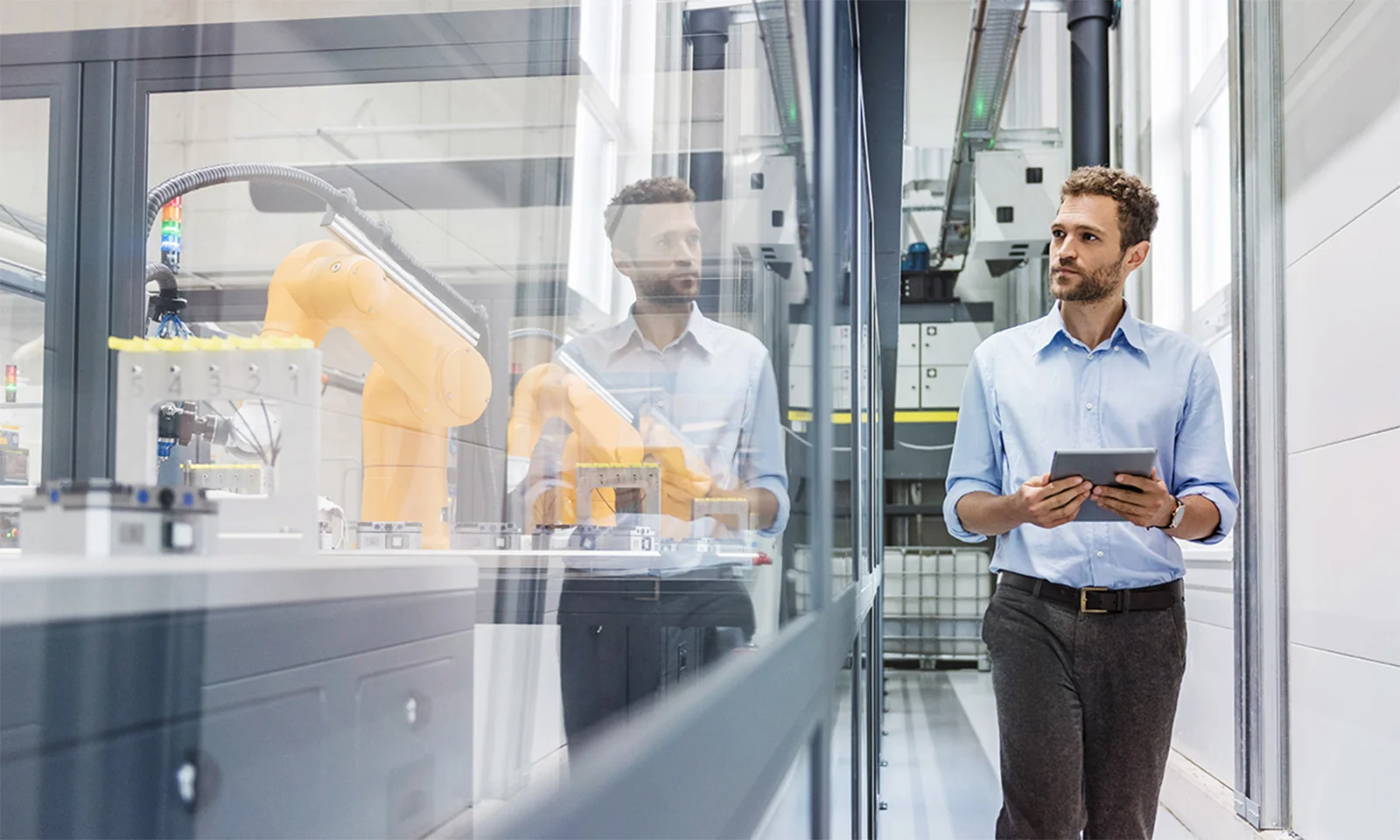 A professional engineer holding a tablet walks through a modern factory with advanced robotic arms working behind glass partitions.