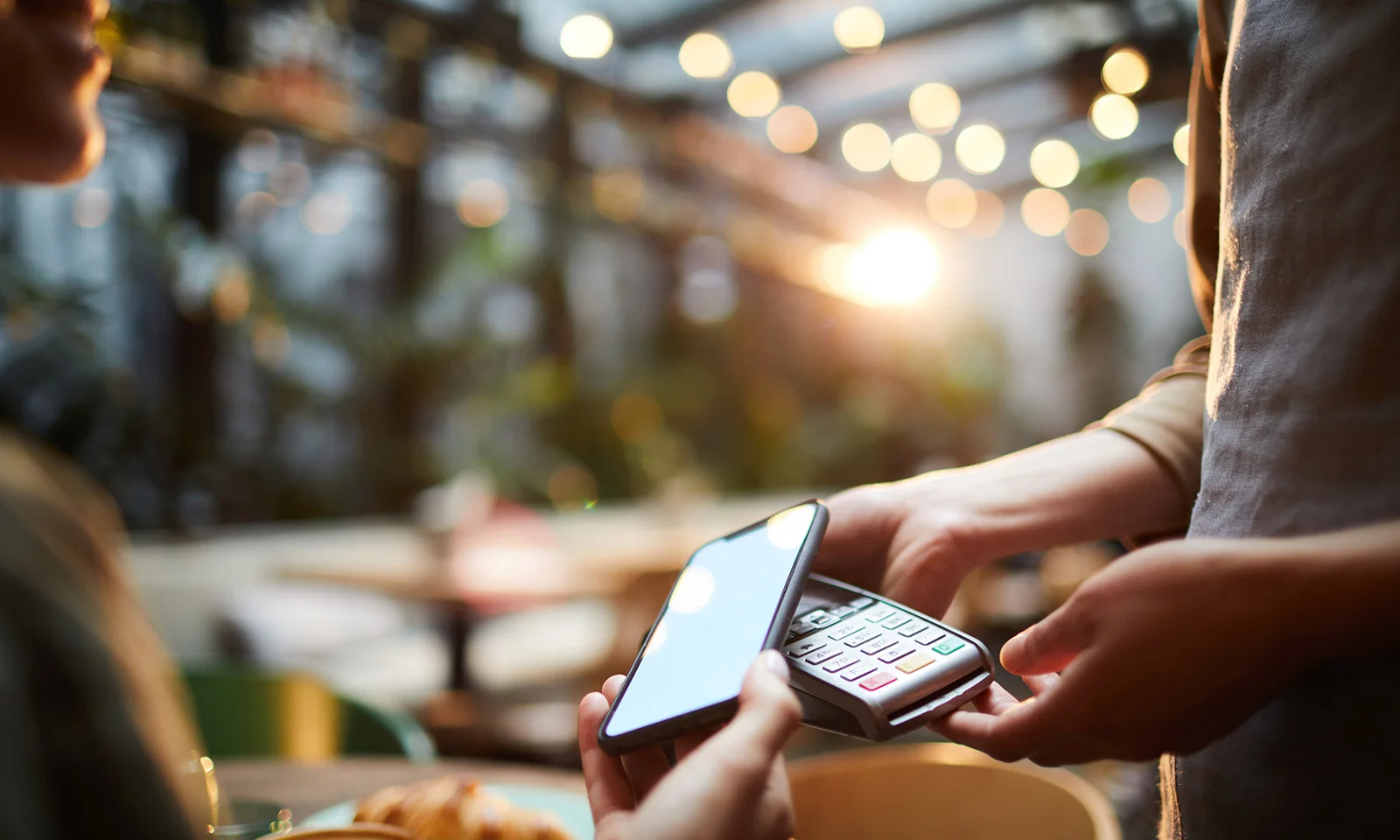 Close-up of a person using a smartphone to make a contactless payment with a card reader in a well-lit café.