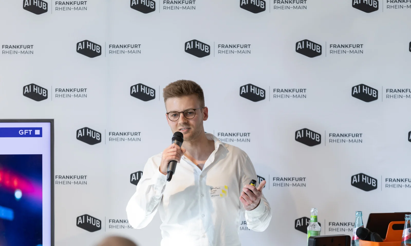 Jonathan Gross von GFT präsentiert das Service Speech Brain auf dem AI Hub Event im TechQuartier Frankfurt. Er steht vor einem Hintergrund mit Logos des AI Hub Frankfurt Rhein-Main und spricht in ein Mikrofon.