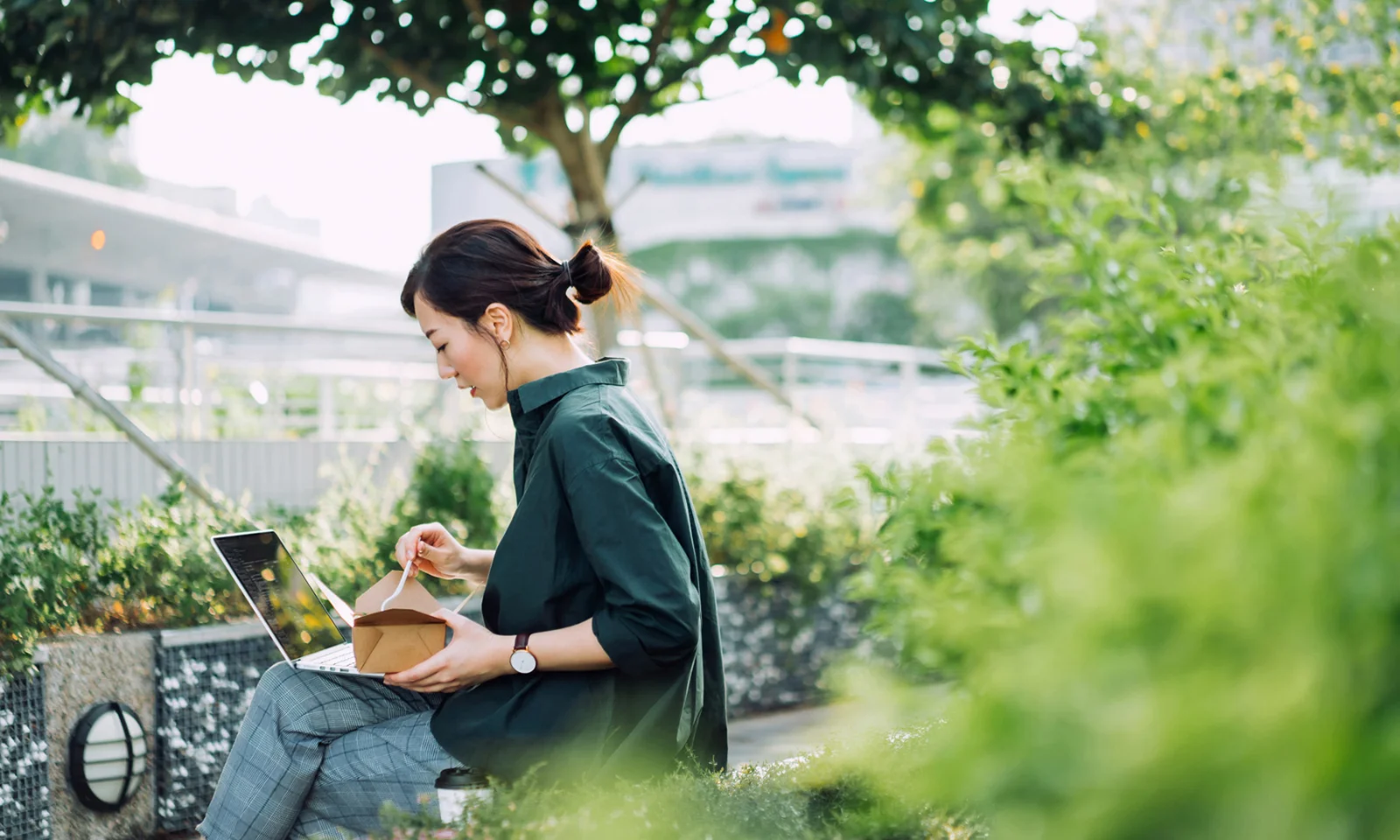 Une femme travaillant sur un ordinateur portable à l&#039;extérieur, entourée de verdure, tout en mangeant dans une boîte à emporter, symbolisant l&#039;intégration de pratiques écologiques dans la vie professionnelle quotidienne et la durabilité.