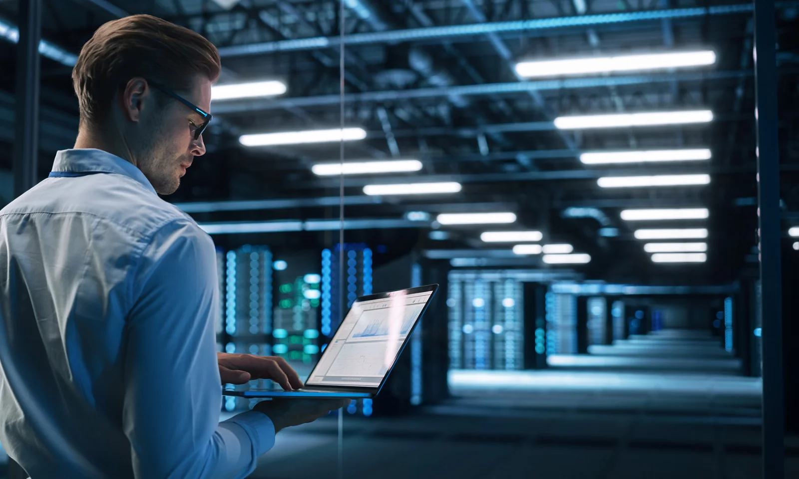 An IT specialist in a white shirt works on a laptop in a modern data center with rows of server racks and bright overhead lights.