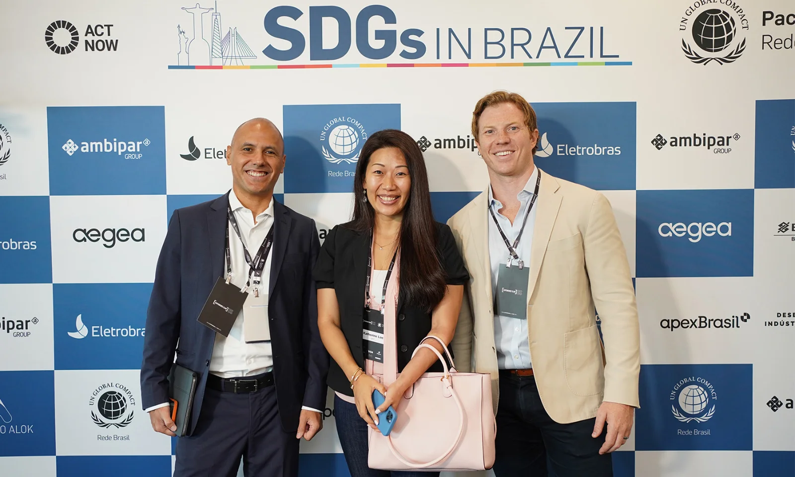 Three attendees smiling for a photo at the SDGs in Brazil event, standing in front of a backdrop featuring sponsor logos including Ambipar, Eletrobras, and ApexBrasil.