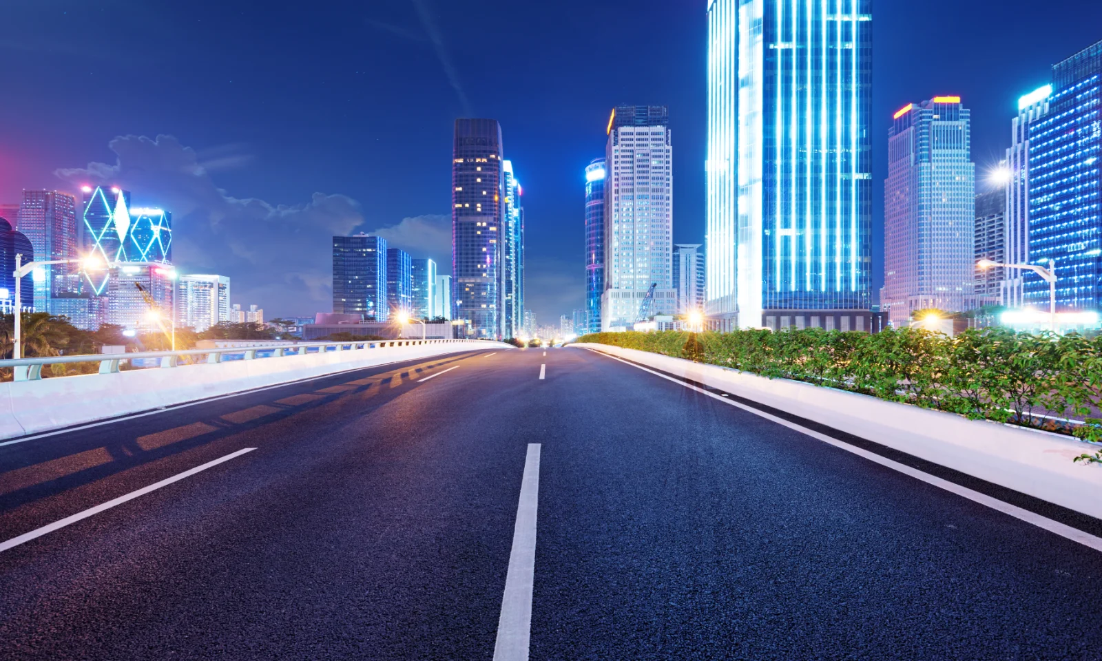 A wide, empty road in a modern city at night, lined with brightly lit skyscrapers, symbolizing urban development and infrastructure.