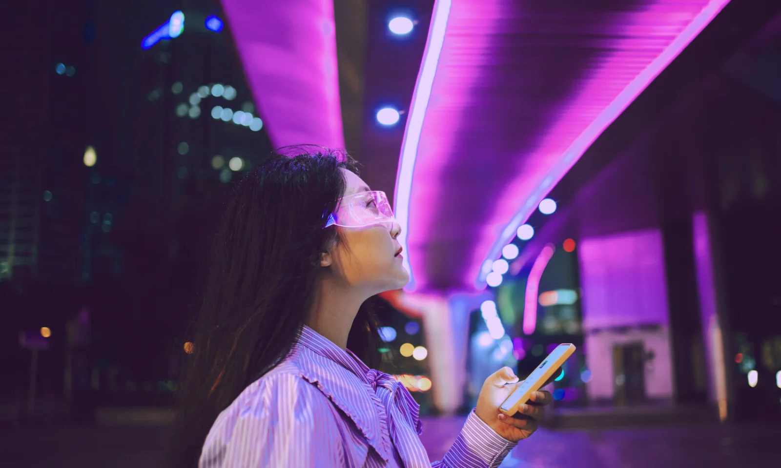 A woman holding a smartphone and looking up at vibrant neon lights on a modern urban bridge at night, symbolizing technology and urban life.