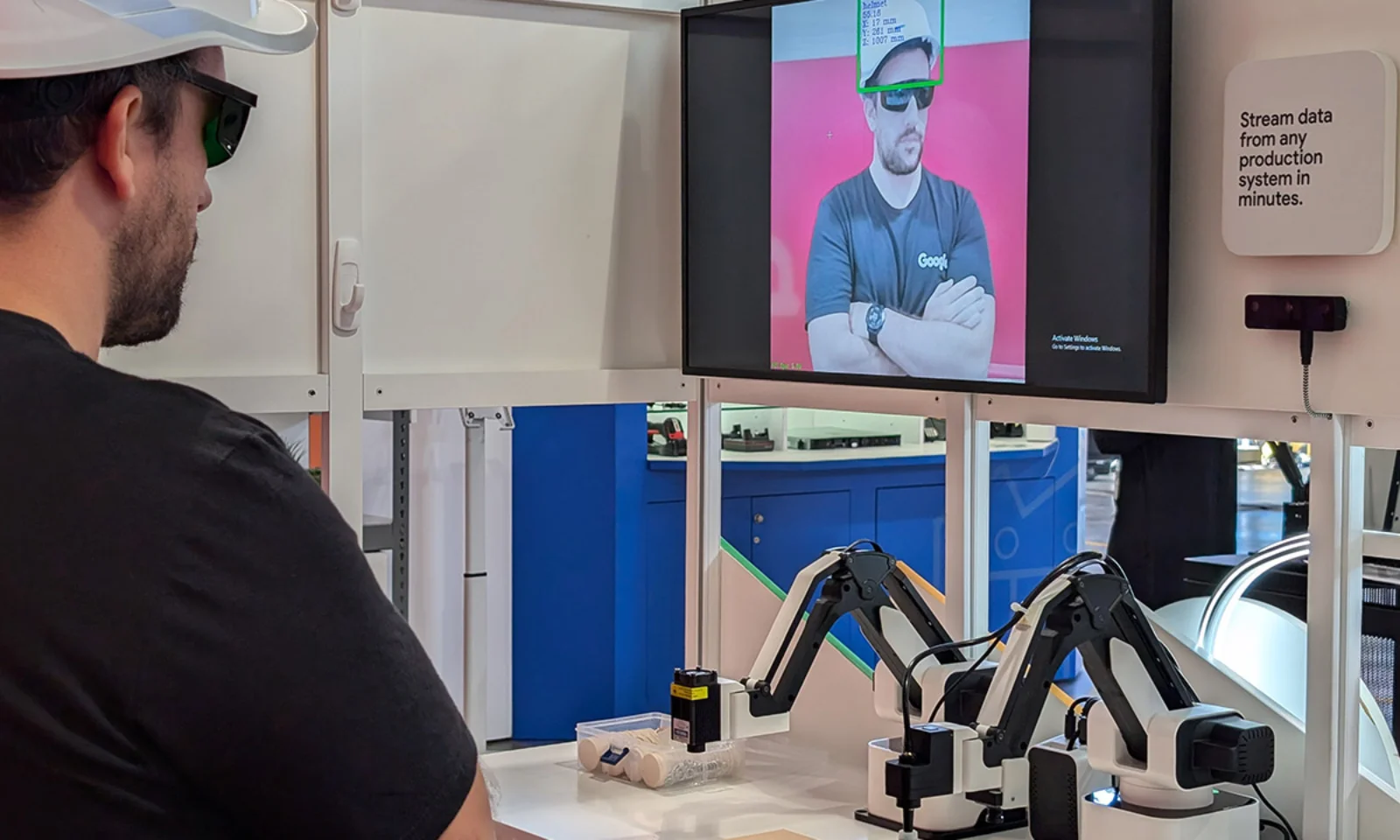 A person in a hard hat and protective glasses interacts with a manufacturing automation system displaying real-time data on a monitor, alongside robotic arms for automated tasks.