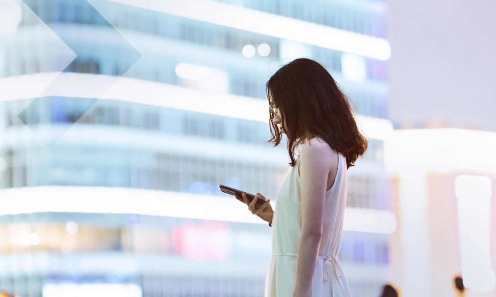 Eine Frau im weißen Kleid steht in einer modernen Stadt und schaut auf ihr Smartphone, umgeben von beleuchteten Gebäuden.