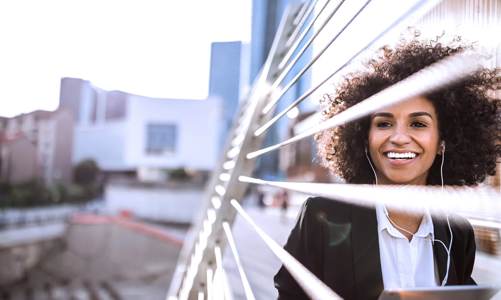 Smiling professional woman with curly hair, listening to music on a modern bridge in an urban setting.