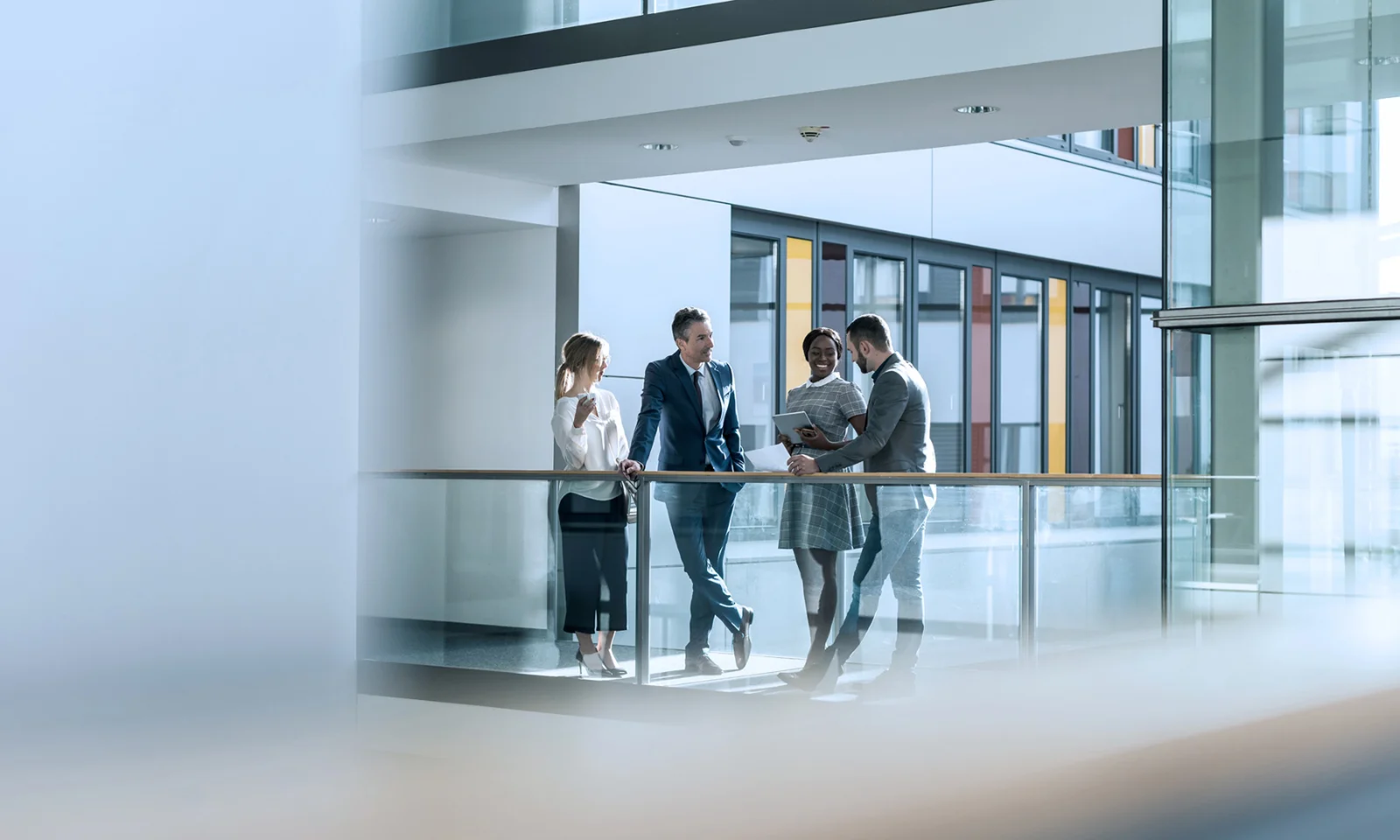 This image captures a professional meeting in a modern office setting. Four individuals, three men and one woman, are engaged in a conversation, standing by a glass railing on an upper floor. The office environment features clean, contemporary design elements with large windows and glass partitions, creating a bright and open atmosphere. The people are dressed in business attire, suggesting a formal and collaborative business environment. This visual is ideal for conveying partnership, collaboration, and a modern business ethos.