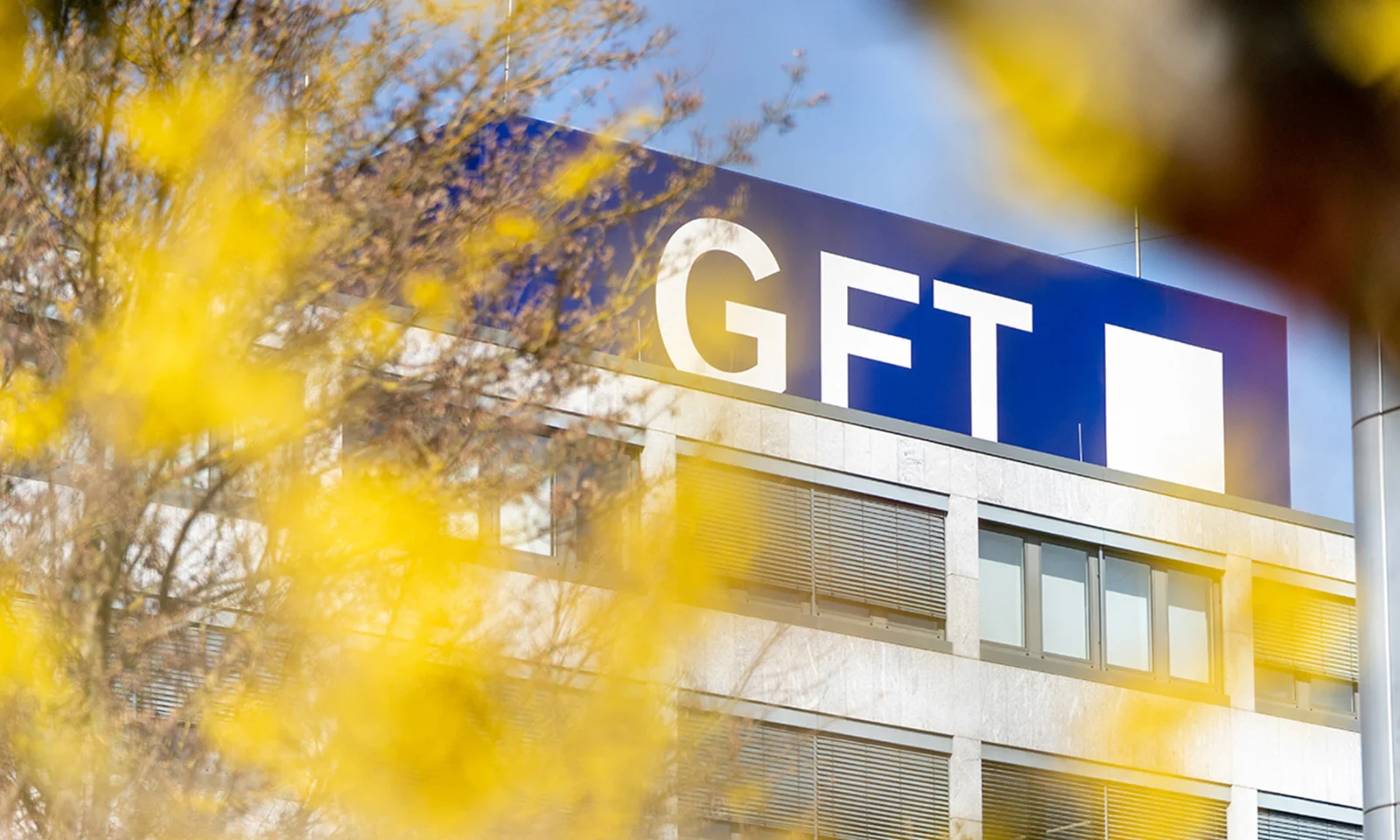 Edificio de la sede de GFT con el logotipo de la empresa destacado en la parte superior, enmarcado por un follaje amarillo vibrante en primer plano.