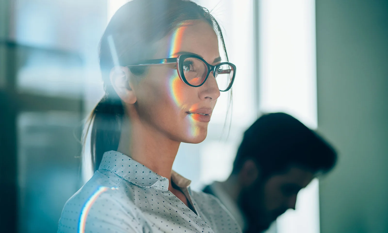 A confident woman wearing glasses looks ahead with a focused expression, with a subtle rainbow reflection on her face, symbolising strategic thinking and modern business acumen.