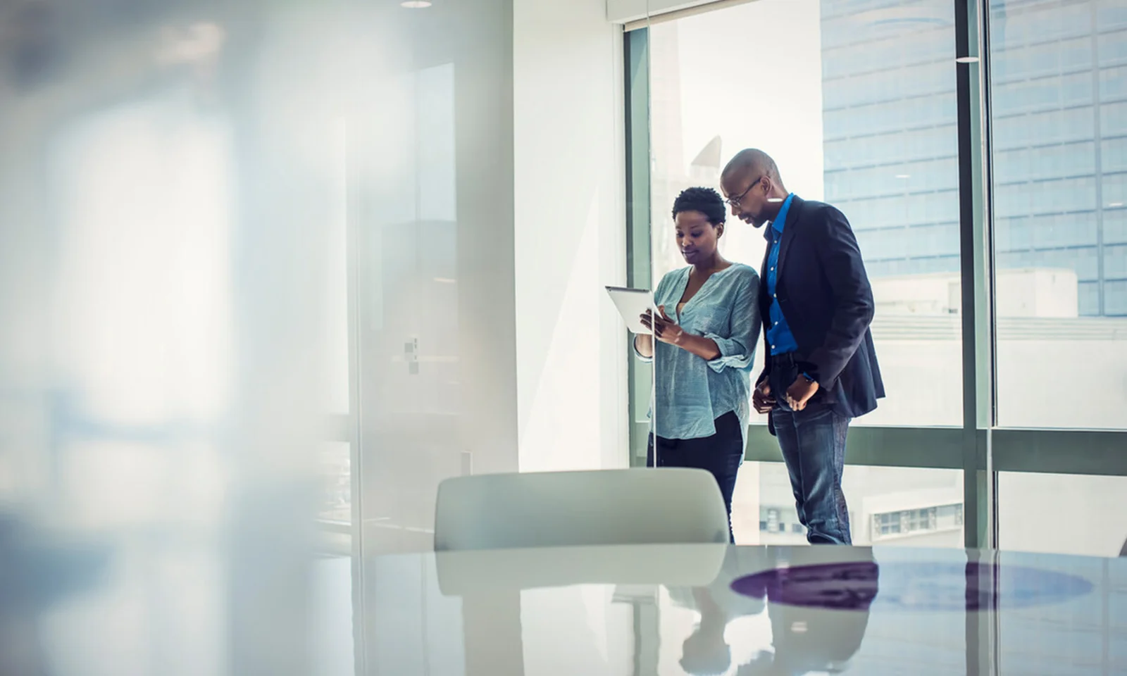 Two business professionals reviewing information on a tablet in a modern, bright office with large windows.