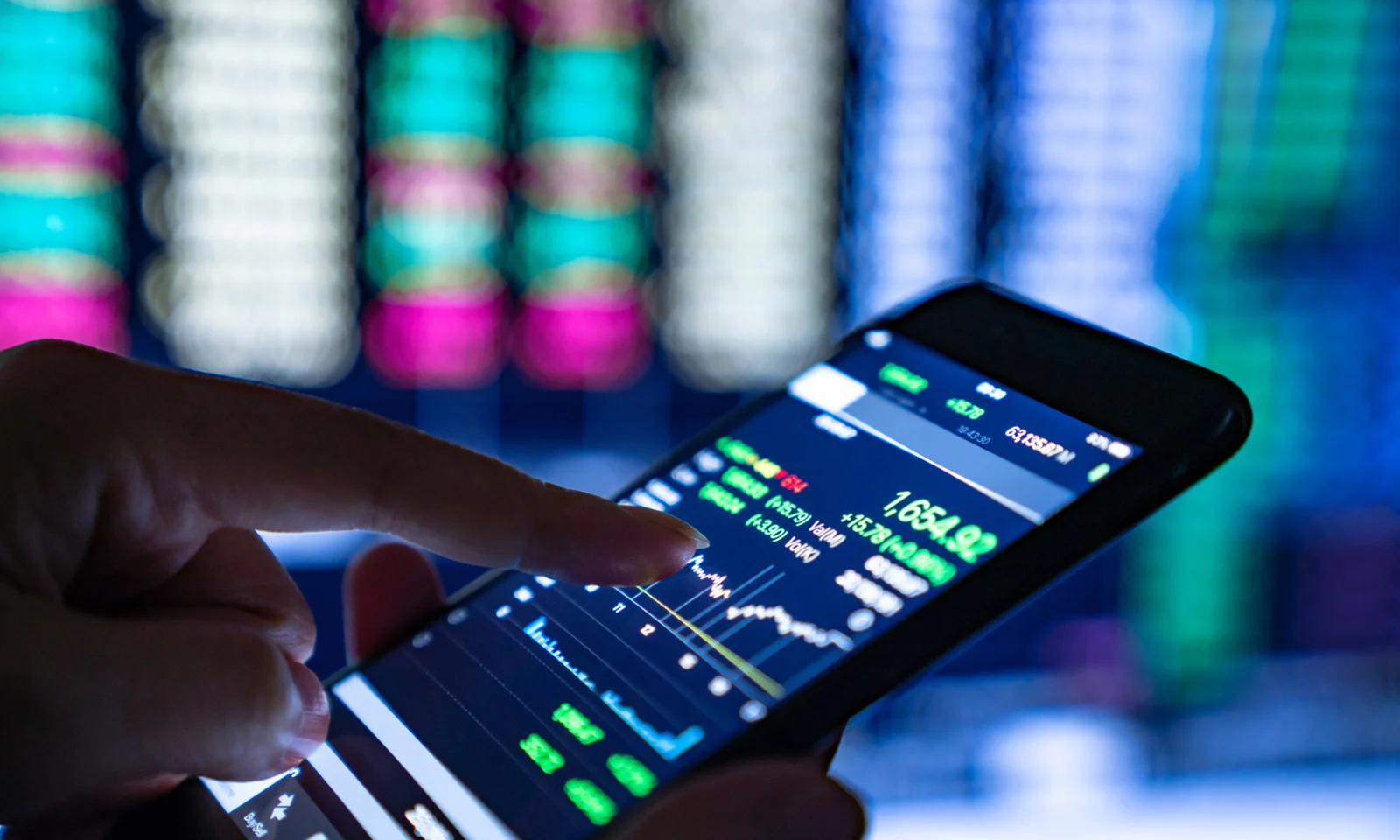 A close-up of a hand using a smartphone to monitor stock market data with blurred financial screens in the background.