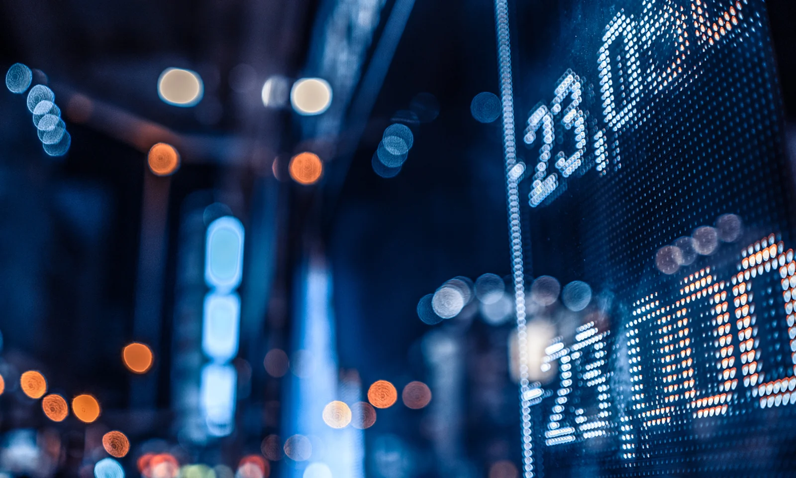 Close-up of a digital stock market display board with blurred city lights in the background, highlighting numbers and financial data.