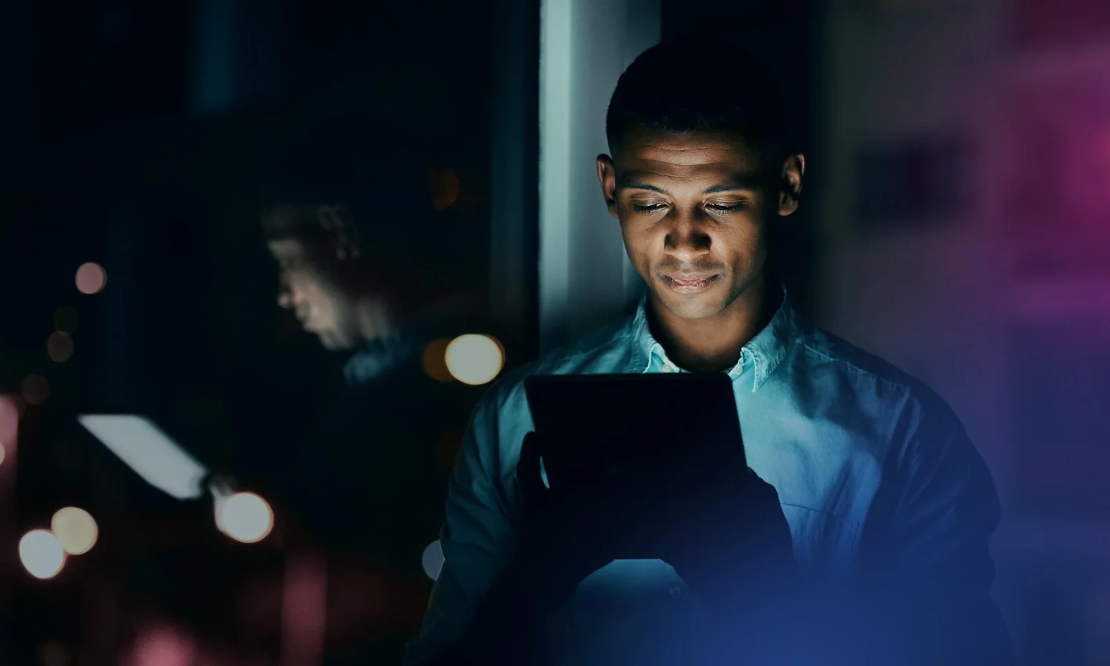 Man looking at a tablet screen at night, standing by a window with city lights reflecting on the glass.