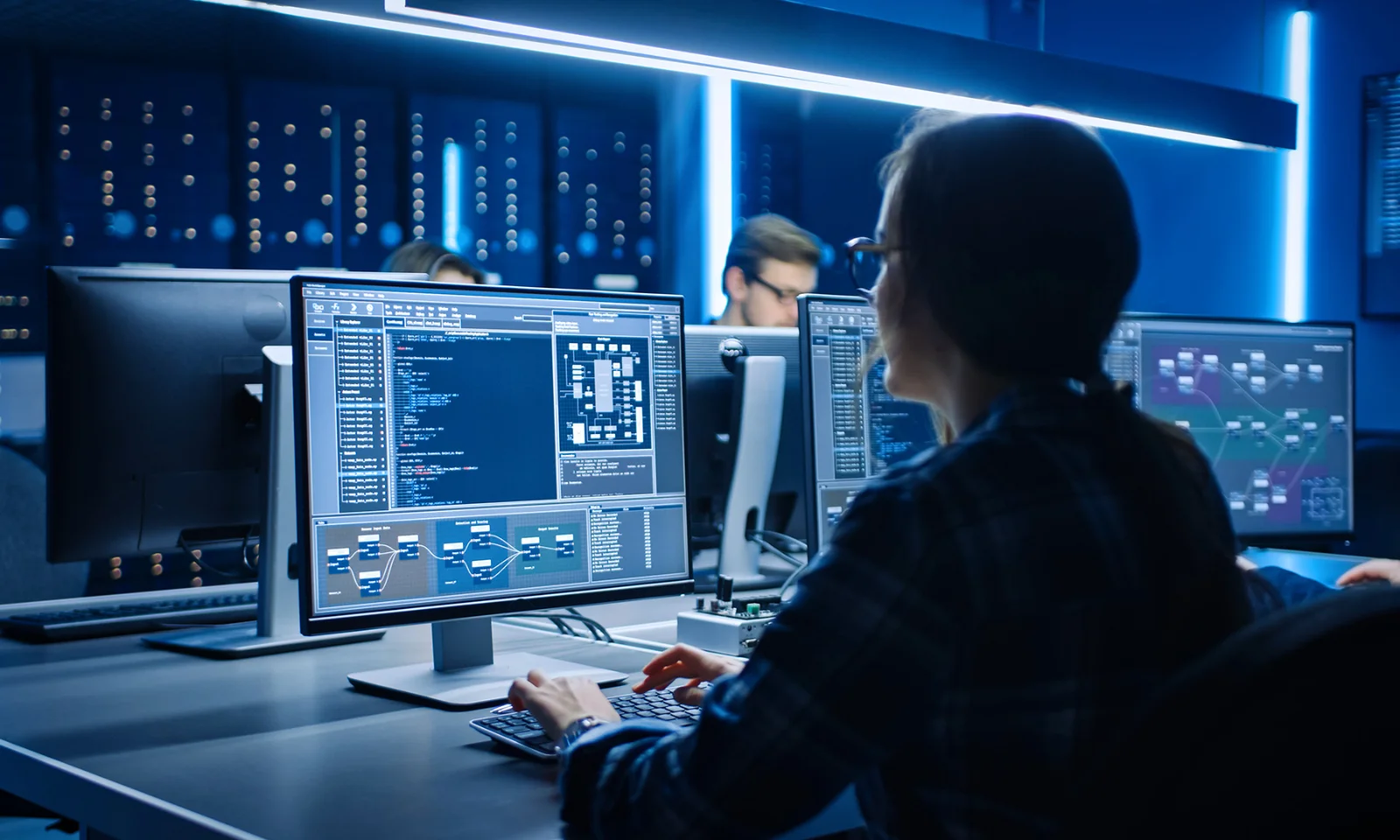 Software engineer working on multiple monitors displaying complex code and data visualisations in a high-tech office environment.