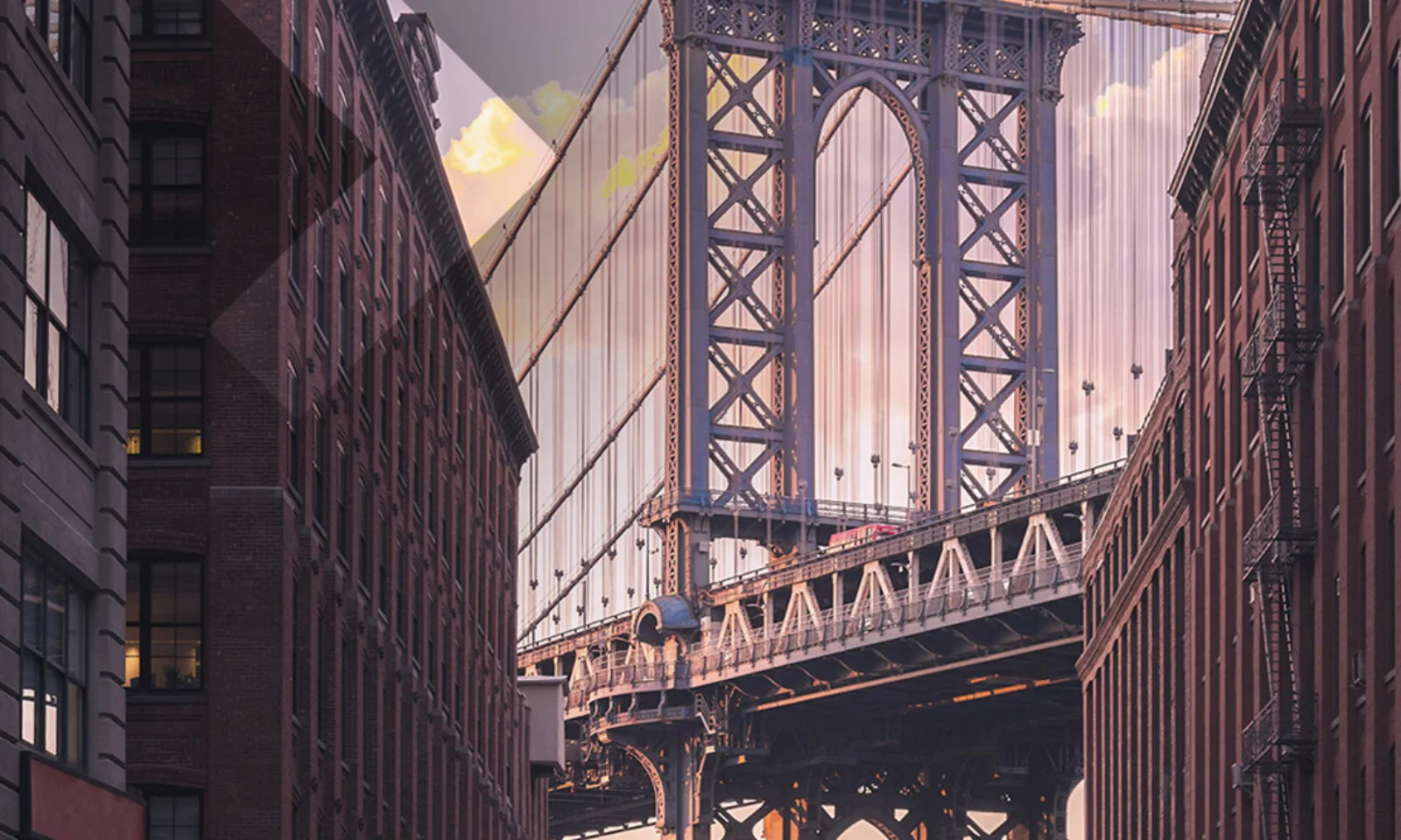 A view of the Manhattan Bridge framed by tall urban buildings at sunset, showcasing the architectural beauty and urban landscape of New York City.