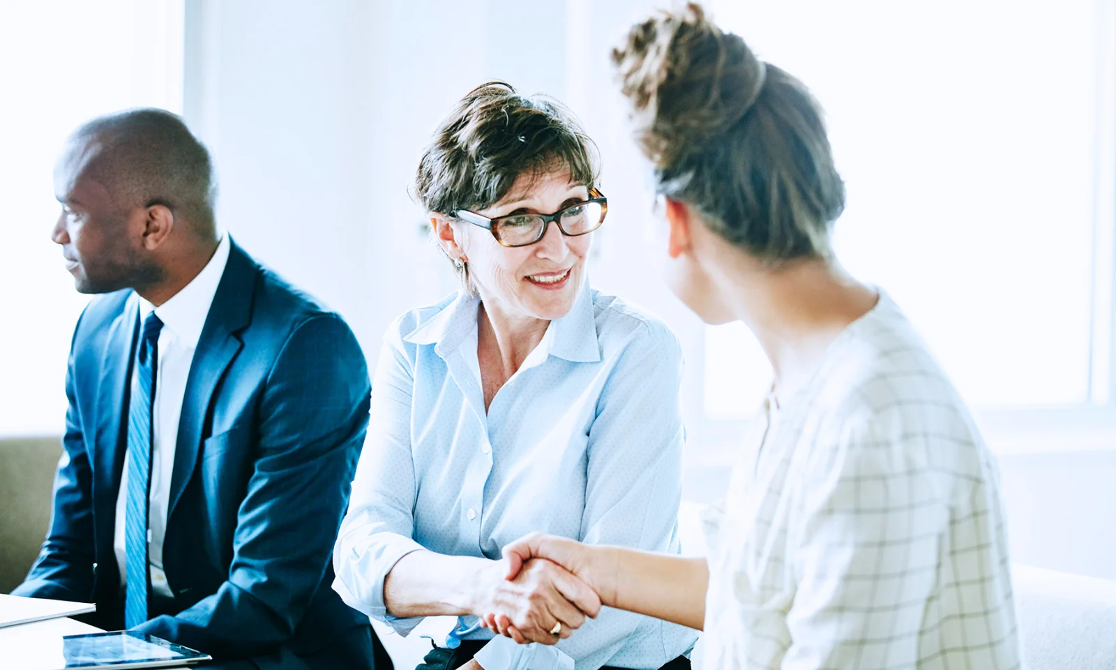 Professionals in a business meeting, with two women smiling and shaking hands, while a man in a suit looks on, symbolising successful networking and collaboration.