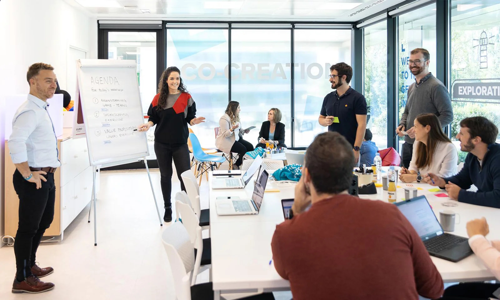 A diverse group of professionals participating in a design services workshop, engaging in discussion and brainstorming, with a flipchart displaying the agenda and laptops on the table.