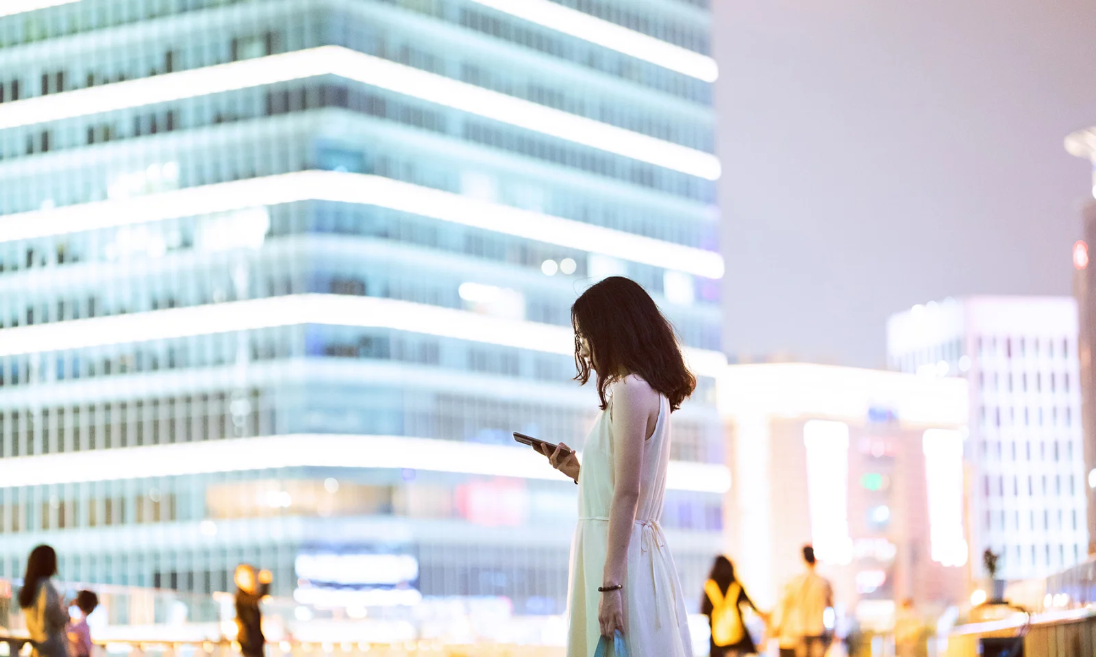 Una mujer parada en un entorno urbano por la noche, mirando su teléfono inteligente, con edificios modernos brillantemente iluminados en el fondo, que representan el tema de la digitalización en la banca.