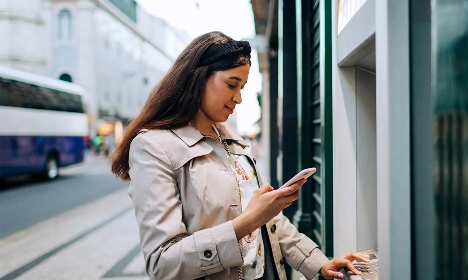 Young woman standing at an ATM while using her mobile phone, showcasing digital banking technology in an urban setting.