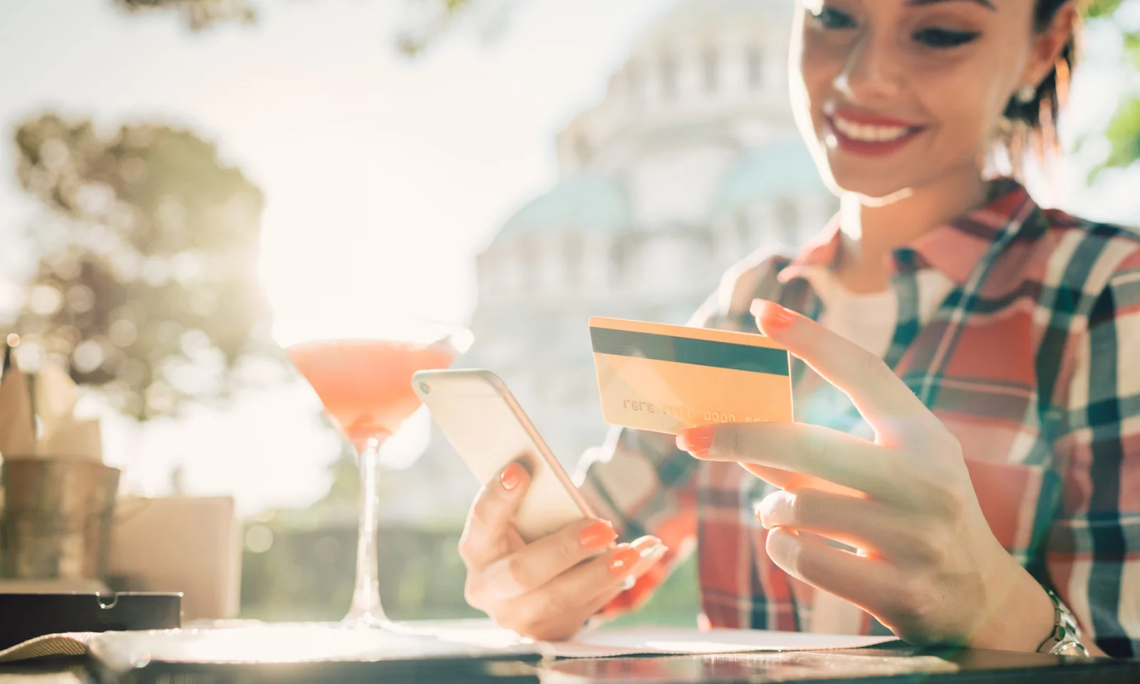 Uma mulher sorridente segurando um smartphone e um cartão de crédito, desfrutando de uma bebida ao ar livre, simbolizando a conveniência e a facilidade dos serviços bancários pessoais modernos.