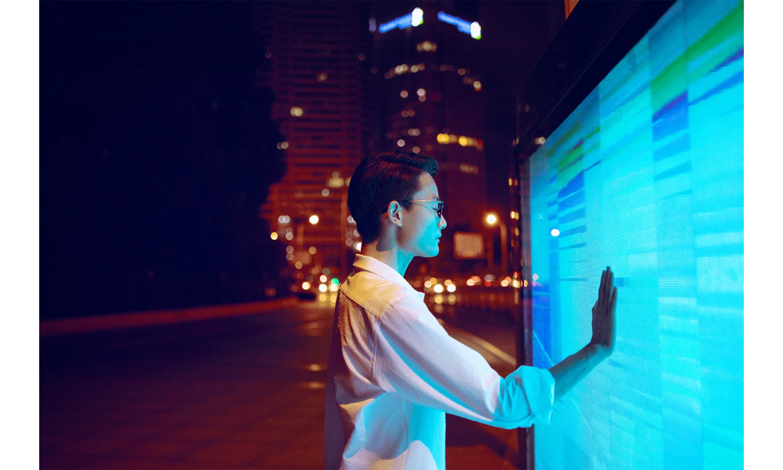 A person in a white shirt interacts with a large, brightly lit digital screen on the street at night, surrounded by the blurred lights of a cityscape. The scene conveys advanced security and monitoring capabilities, symbolizing AWS GuardDuty.