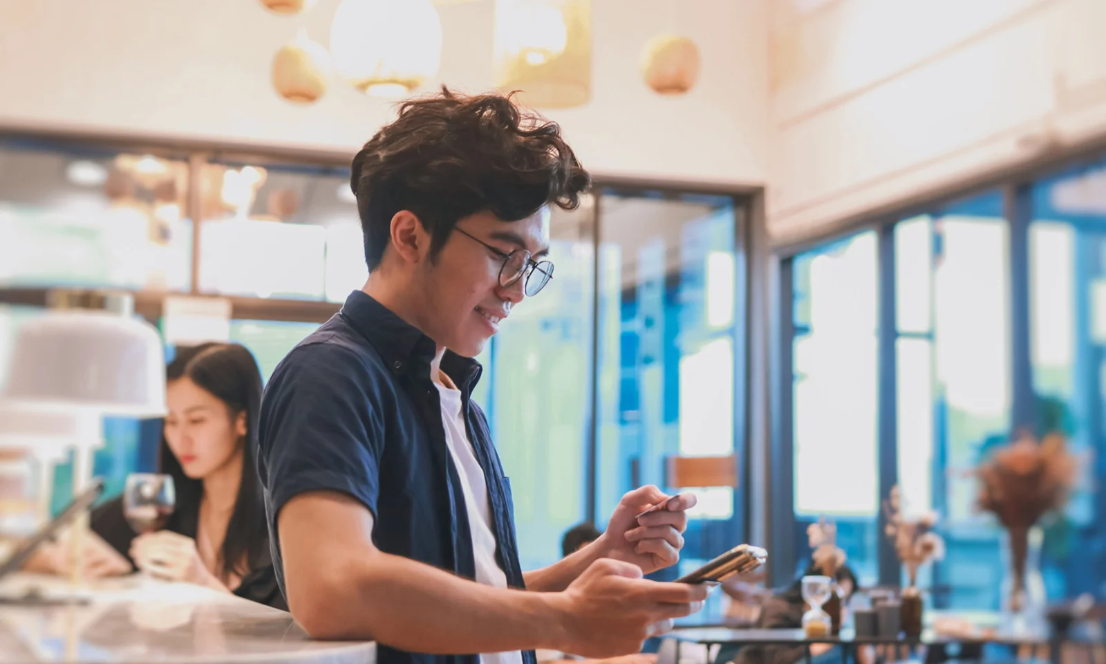 A young man using his smartphone and holding a credit card in a modern café, representing the use of a mobile push notifications platform by a retail bank.