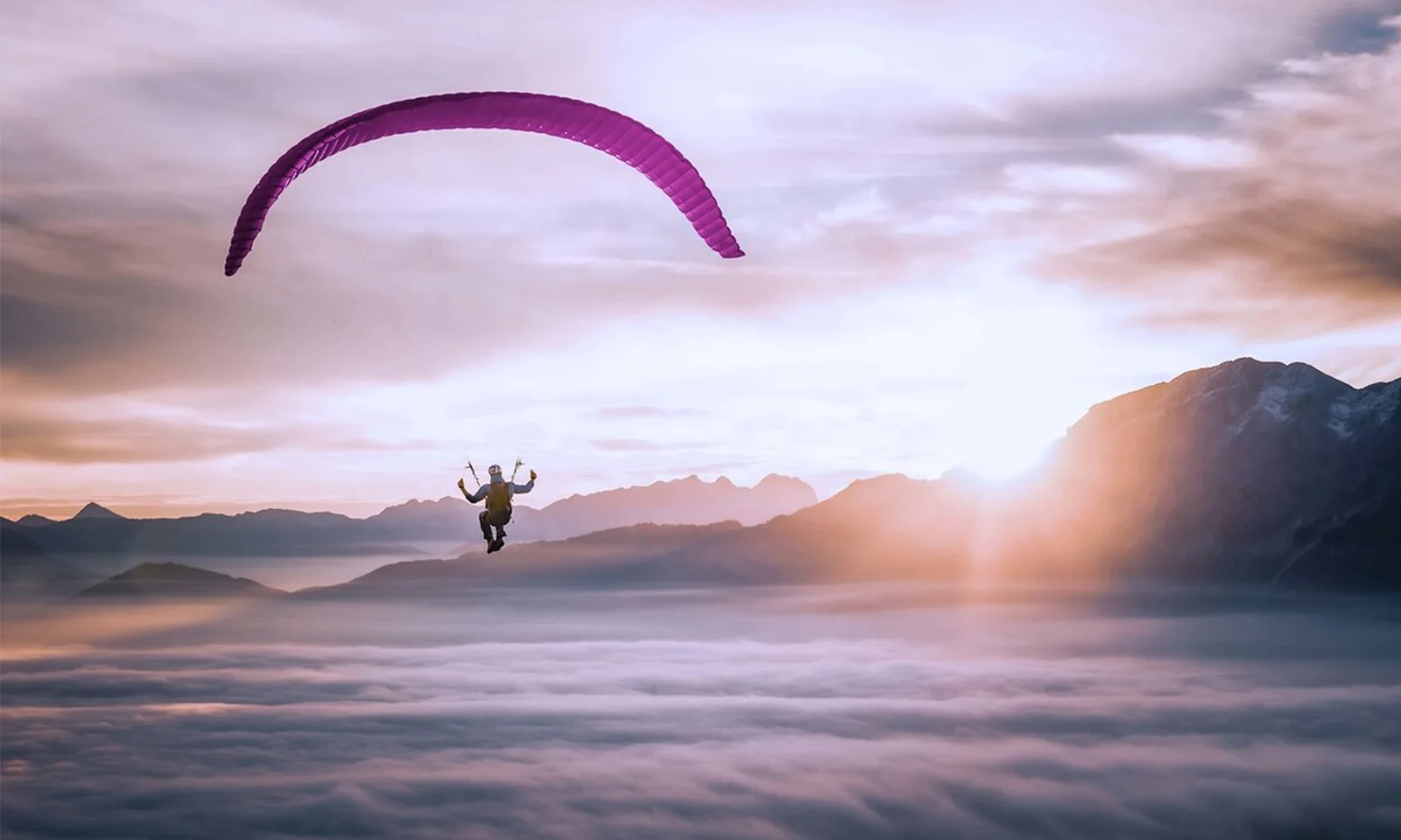 Esta imagem de tirar o fôlego captura a essência da liberdade e da aventura enquanto um parapente solitário voa graciosamente acima de um manto de nuvens. O parapente, com um dossel roxo vibrante, está posicionado contra o cenário deslumbrante de um nascer do sol, lançando um brilho dourado e quente sobre a cena. Os picos das montanhas distantes se elevam acima da camada de nuvens, adicionando profundidade e uma sensação de escala à imagem. A atmosfera serena e inspiradora destaca a beleza do parapente e a perspectiva única que ele oferece sobre o mundo. Este visual encapsula perfeitamente a emoção e a tranquilidade de voar em céu aberto.