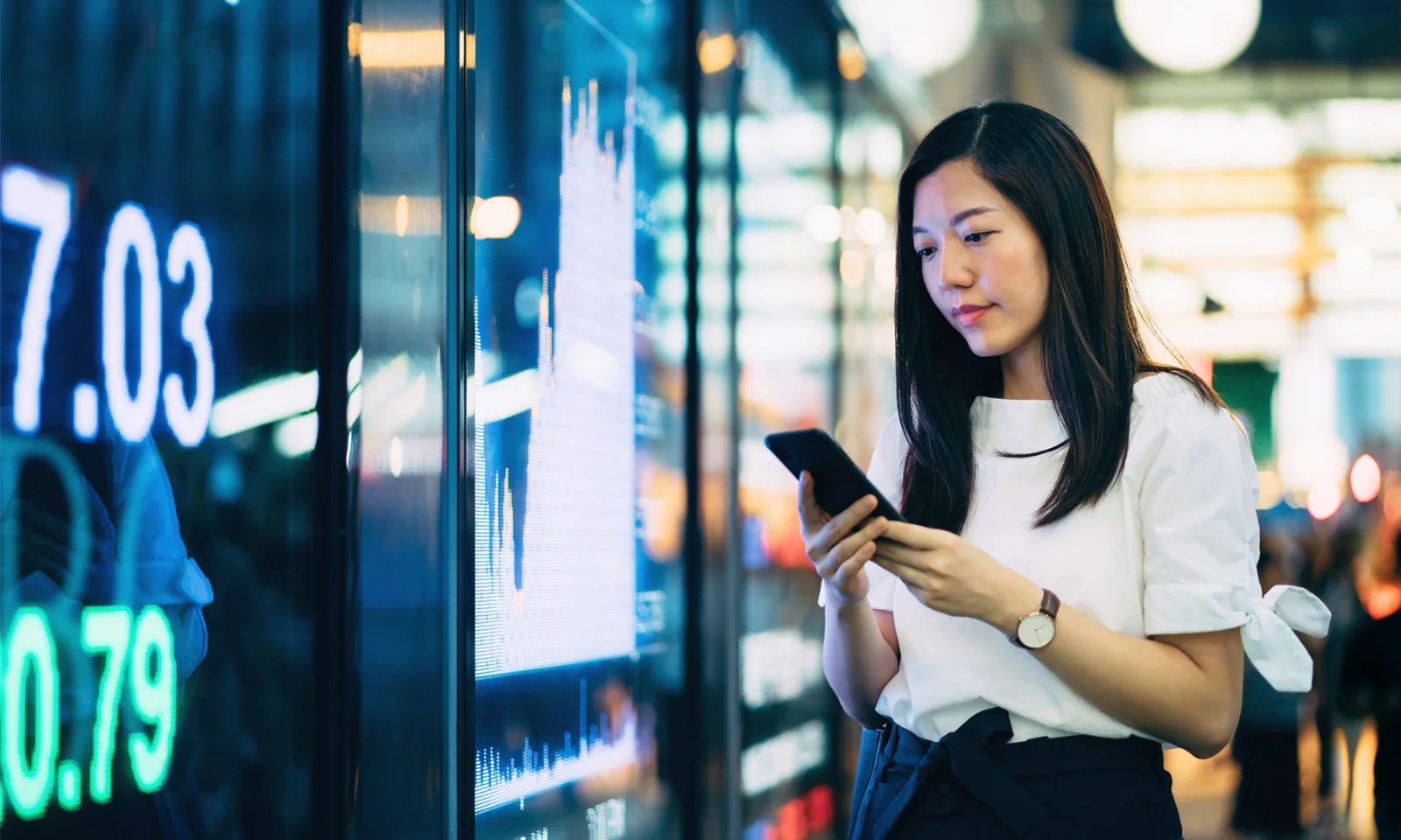 A woman standing in front of an electronic board displaying financial data, using her smartphone, representing the concept of sustainability in technology and business practices.