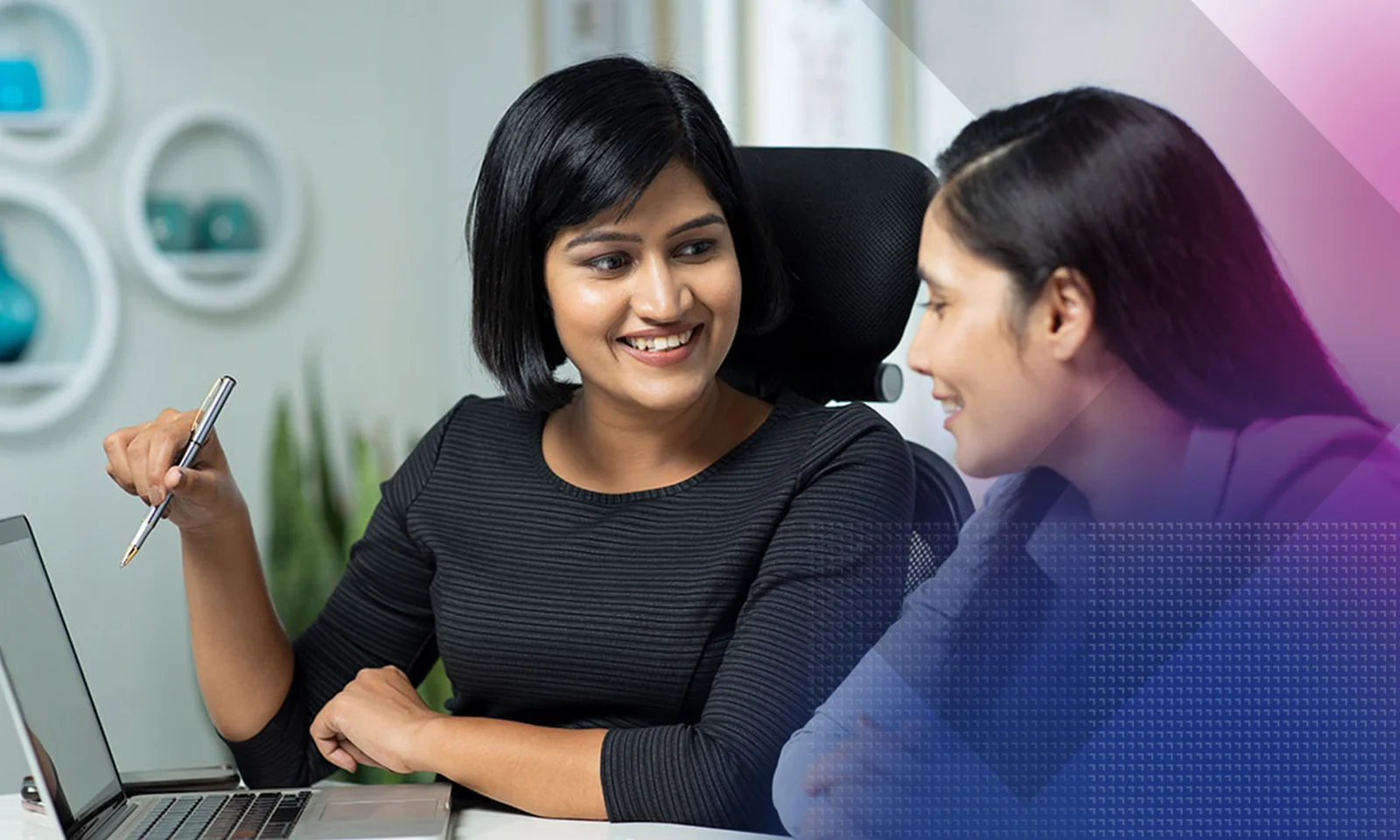 Two professional women engaged in a discussion at a desk, with a laptop open and one holding a pen, in a modern office environment.