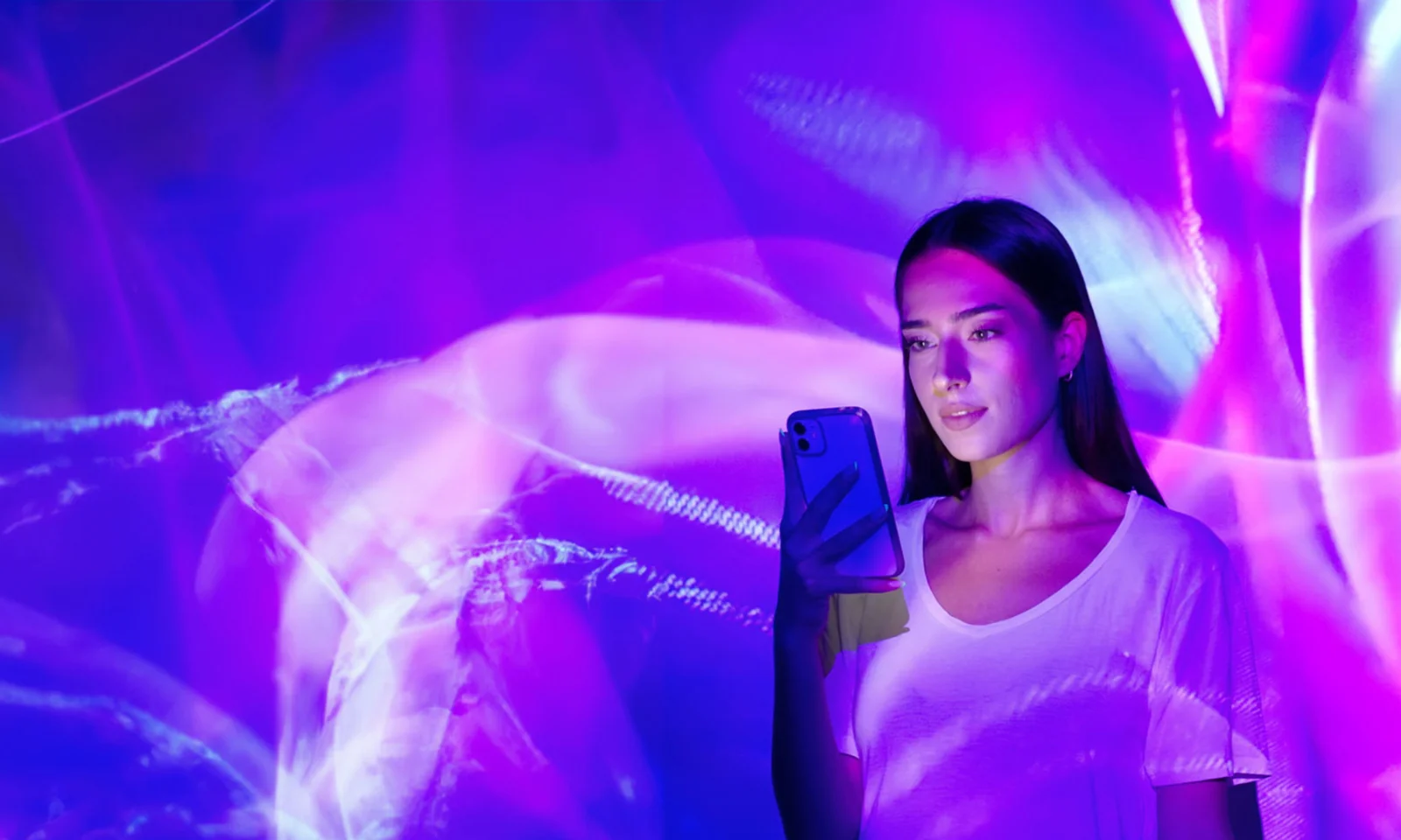 Une femme tenant un smartphone se tient devant un écran lumineux violet et rose vibrant et abstrait.