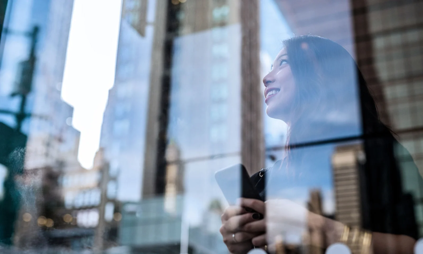 A woman holding a smartphone, smiling while looking up, with the reflection of skyscrapers on a glass window, symbolizing innovation and growth.