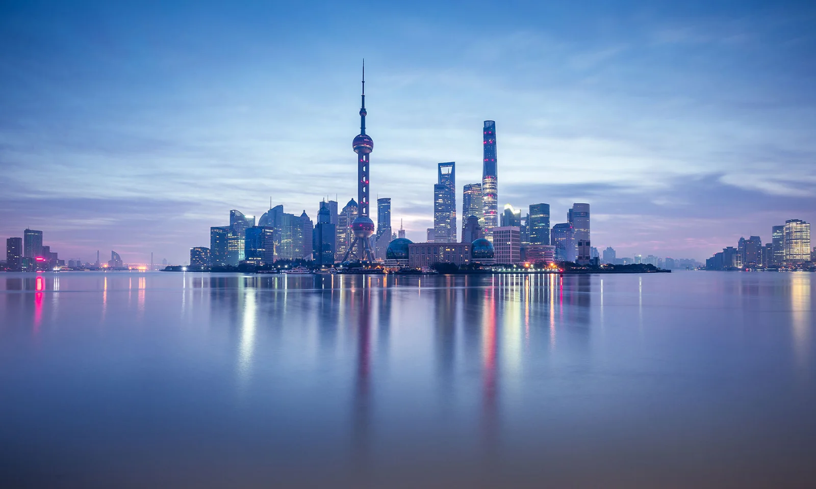 Uma vista panorâmica do horizonte de Xangai ao anoitecer, com arranha-céus modernos e a icônica Torre Pérola Oriental, refletida nas águas calmas do Rio Huangpu.