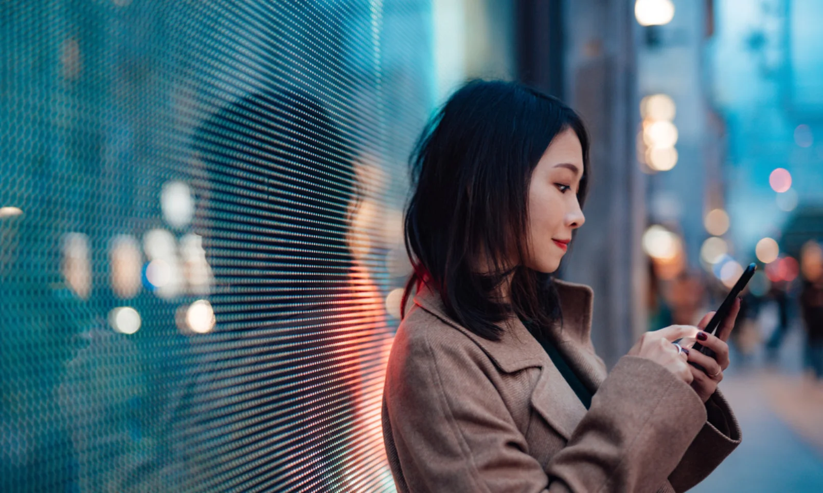 Une femme se tient debout devant un écran numérique la nuit, en train d&#039;utiliser son smartphone. La lumière de l&#039;écran se reflète sur son visage et sur les alentours, créant une ambiance moderne et technophile.