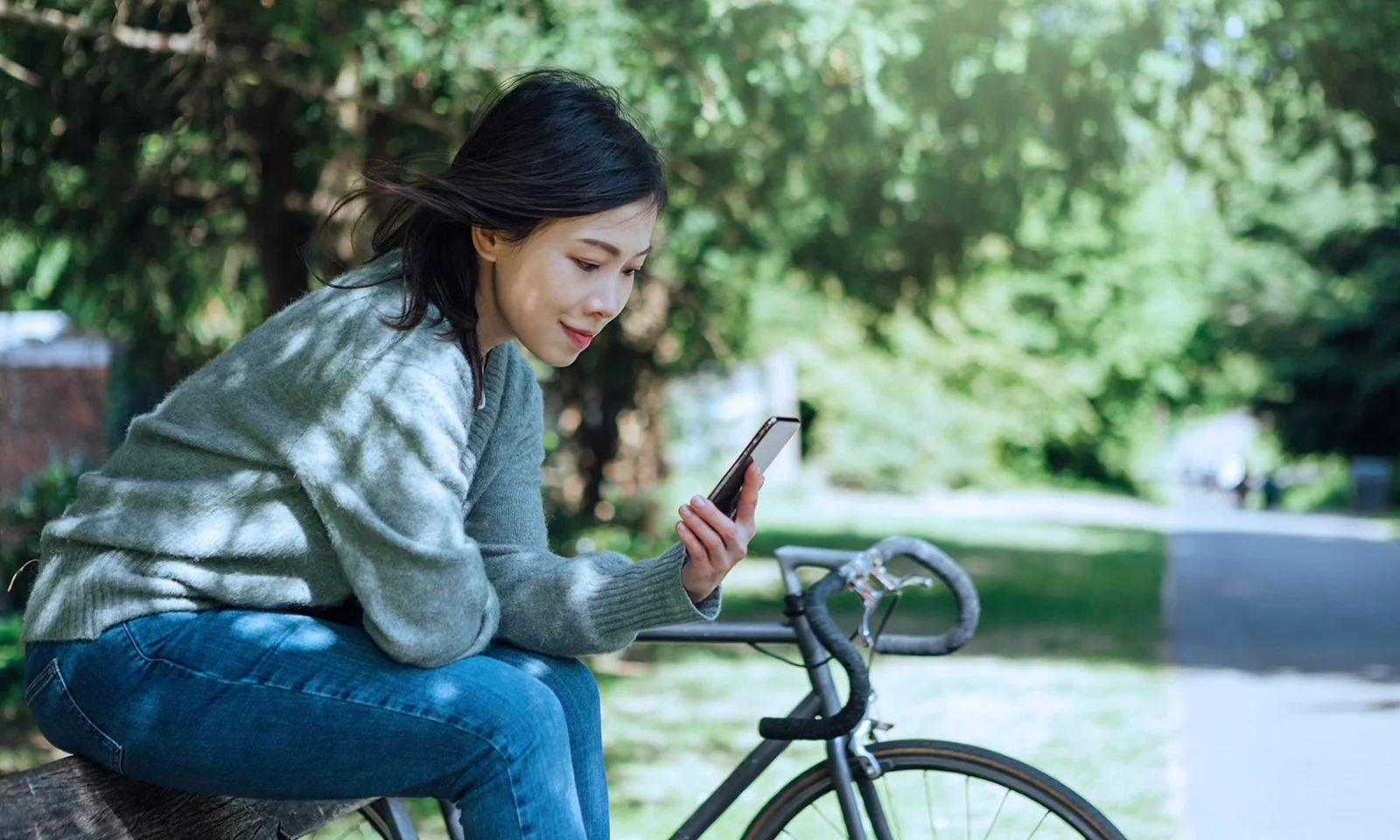 A woman sitting on a bicycle, looking at her phone, in a green park setting, representing sustainability by design