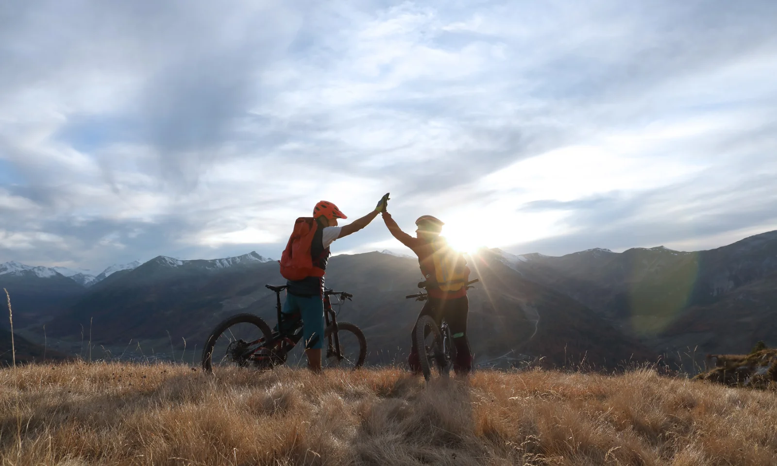 Two cyclists giving a high-five on a mountain top at sunrise, symbolising partnership and achievement.