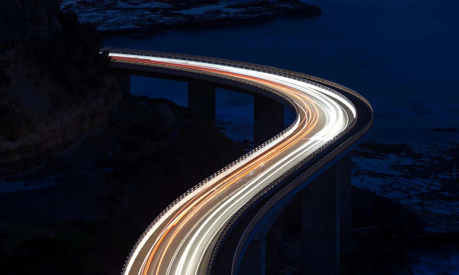 Long-exposure photograph capturing streaks of light from vehicles traveling on a curved bridge over water at night, symbolizing speed and modern infrastructure.