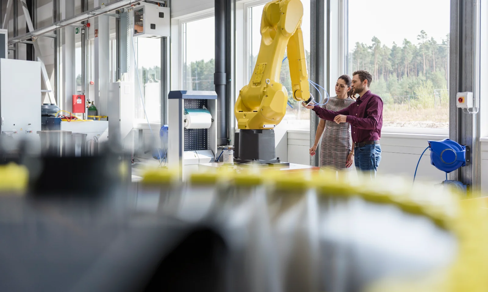 Mann und Frau betrachten einen Roboterarm in einer Fabrikhalle, im Hintergrund ist ein Wald zu sehen. Key Visual für Industry Solutions.