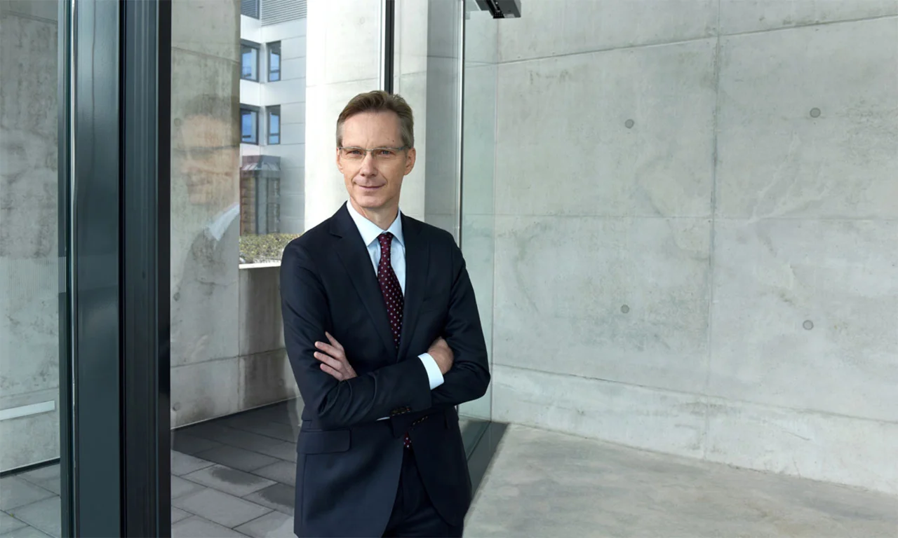 Jochen Rütz, Member of the Administrative Board, standing with arms crossed, reflecting by a glass window in a modern building