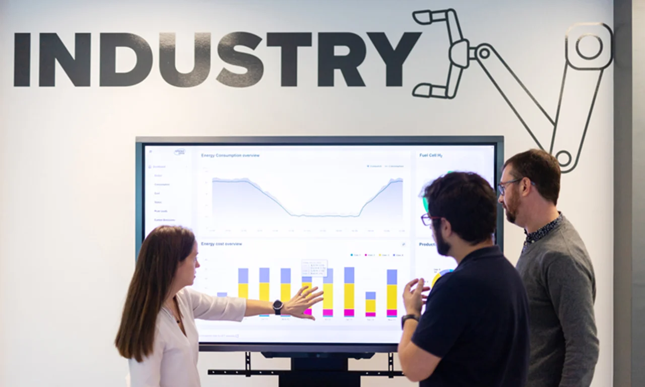 Three professionals discussing data on a large screen in an industry-themed innovation lab space.